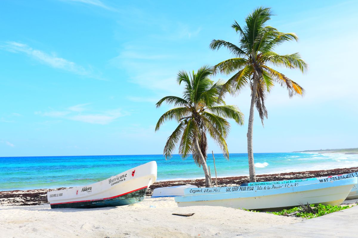 Pláž na ostrově Cozumel s&nbsp;dvěma rybářskými loděmi na písku a&nbsp;palmami pod modrou oblohou.