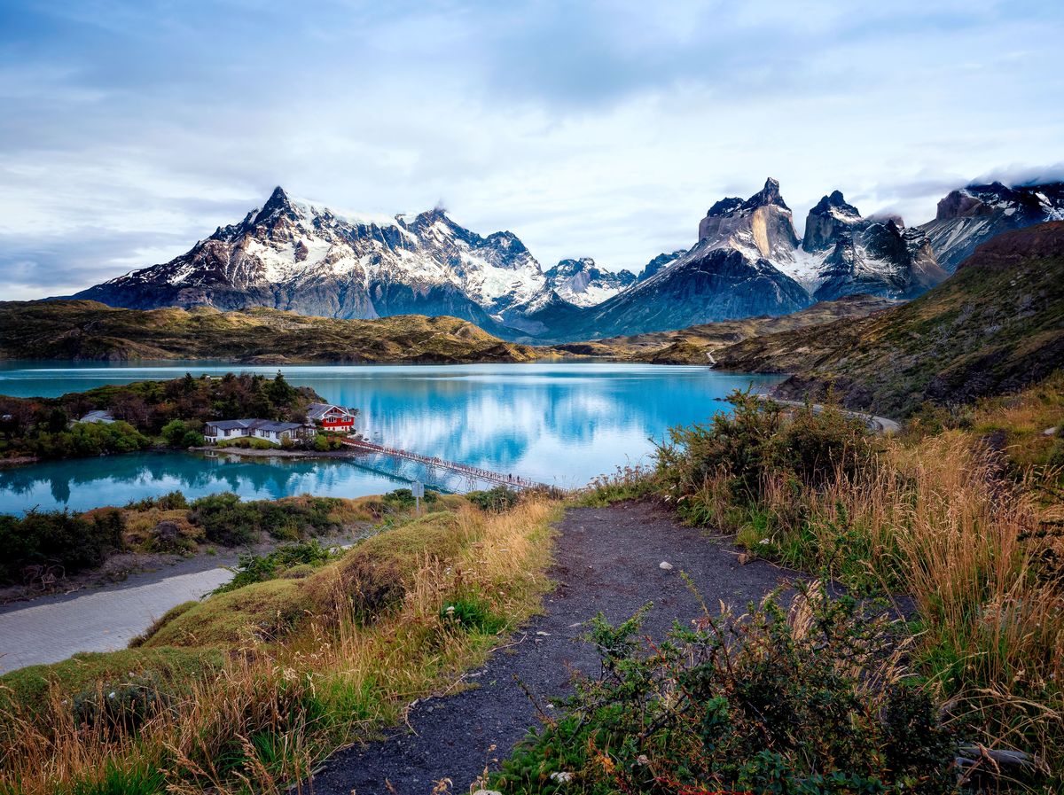 Křišťálově čisté jezero uprostřed chilského národního parku Torres del Paine je obklopené majestátními zasněženými horami a&nbsp;zelení s&nbsp;červenou chatou na pobřeží.