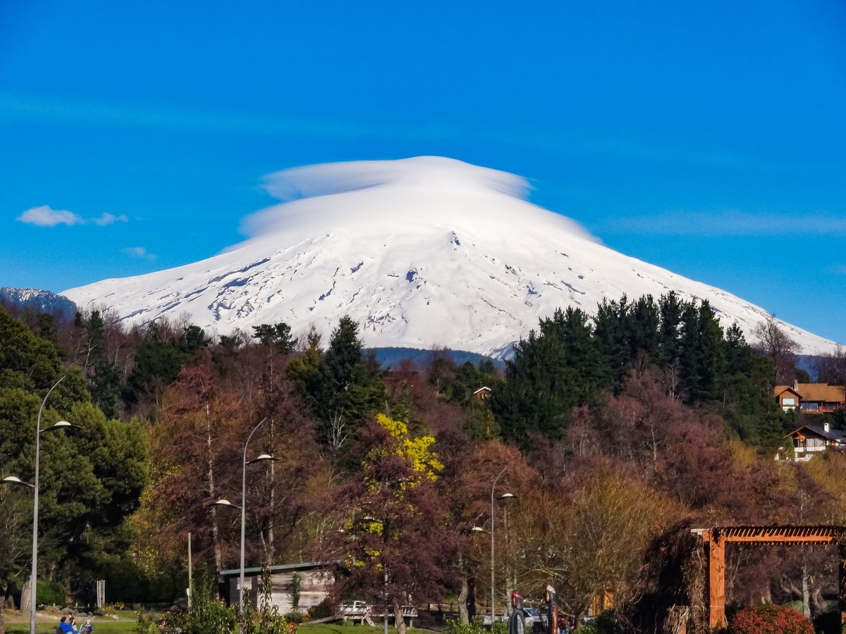 Snímek zasněžené sopky Villarrica v&nbsp;Chile s&nbsp;typickým mrakem kolem vrcholu a&nbsp;lesem v&nbsp;popředí.