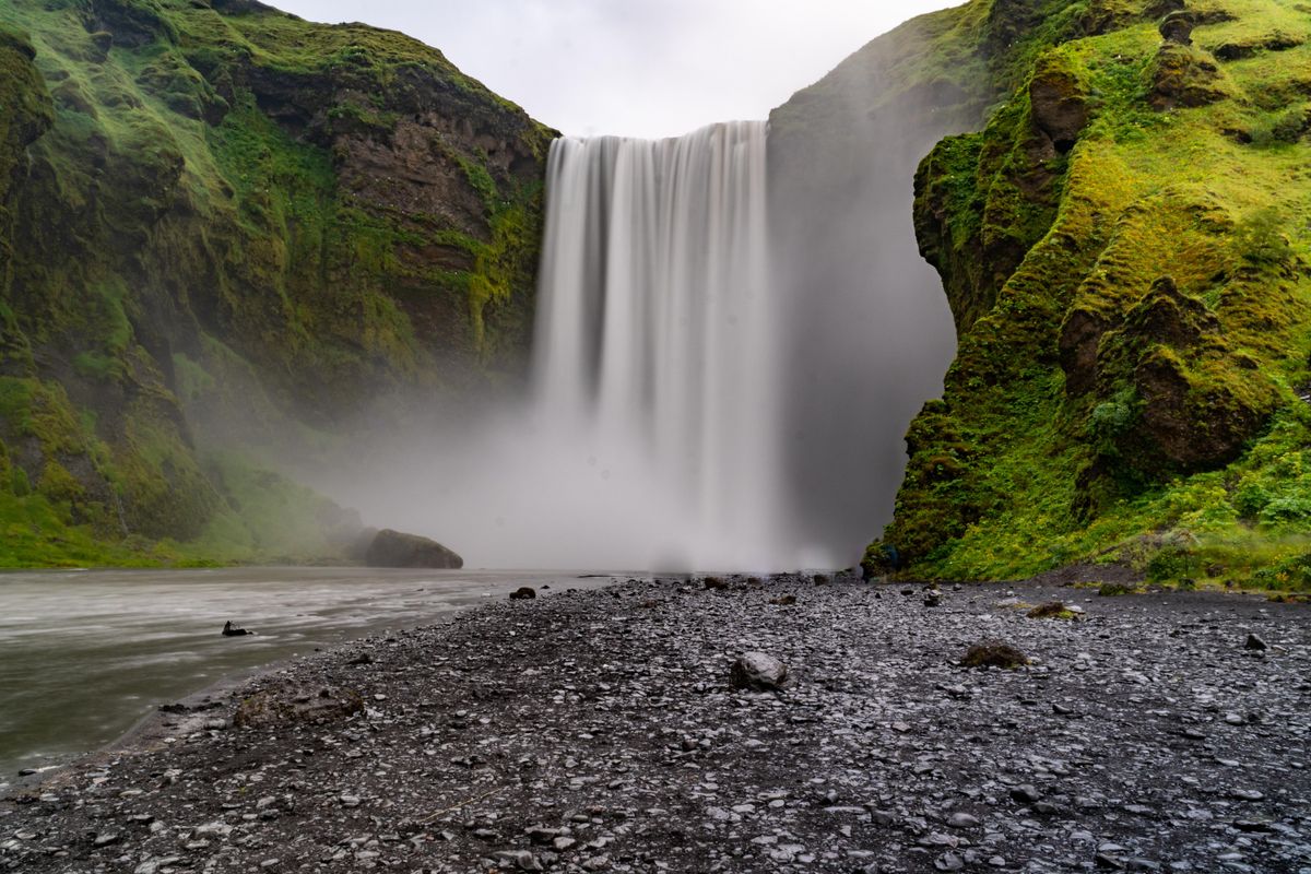 Mohutný vodopád Skógafoss na Islandu, obklopený zelenými svahy a kamenným břehem pod ním.