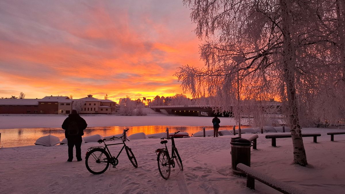 Zimní krajina v&nbsp;Oulu, kde se dvě kola nacházejí na zasněženém břehu řeky pod oranžovou oblohou při západu slunce, zatímco lidé pozorují panoráma přes zamrzlý most.