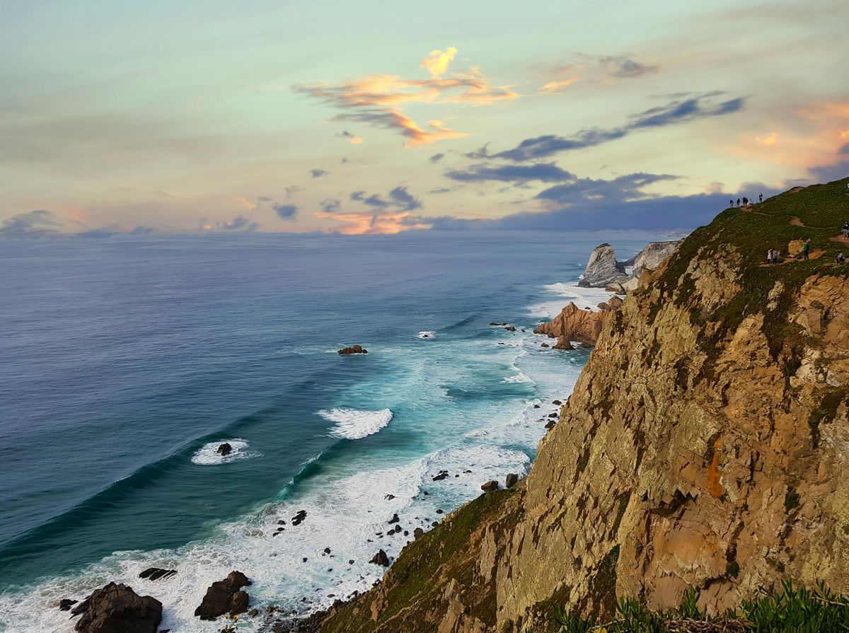 Pobřežní útesy s vlnami Atlantického oceánu a výhledem na mys Cabo da Roca při západu slunce v pozadí.