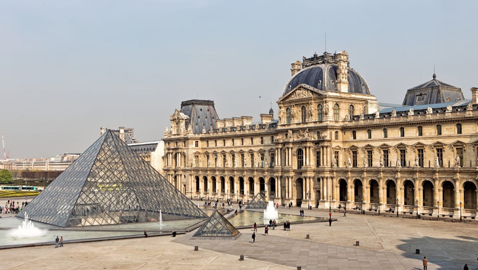 Skleněná pyramida a historická budova muzea Louvre v Paříži s lidmi procházejícími kolem fontán.