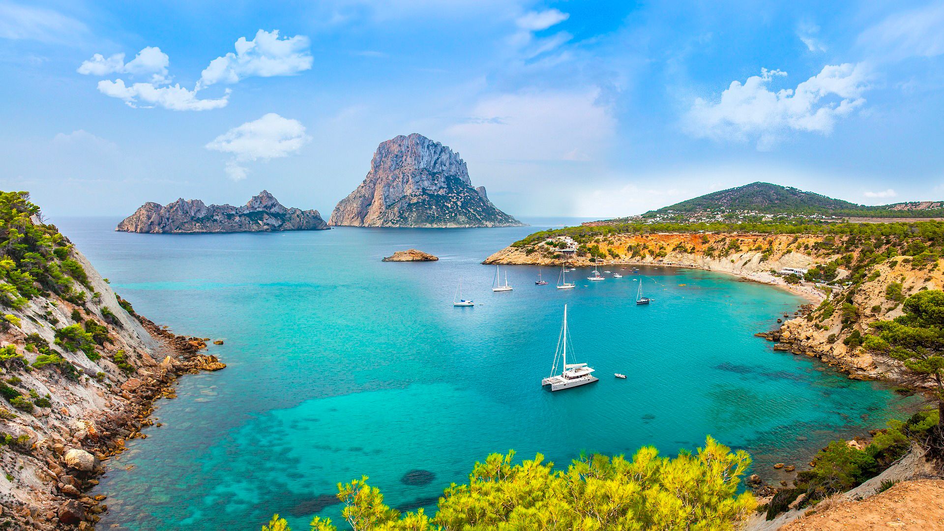 Krásný výhled na zátoku Cala d'Hort na ostrově Ibiza, kde v tyrkysovém moři kotví několik lodí a v pozadí se tyčí dramatické skalní útesy Es Vedrà.