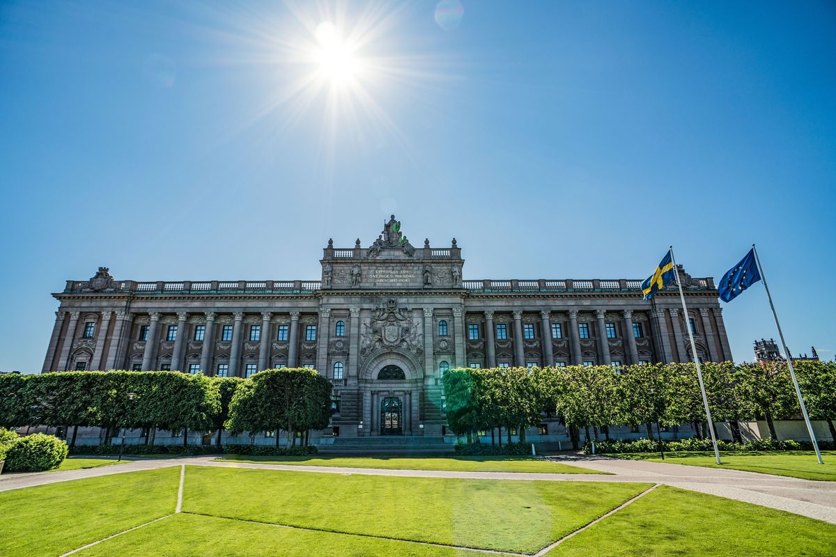 Budova švédského parlamentu Riksdag ve Stockholmu pod jasnou modrou oblohou se dvěma vlajkami v popředí.