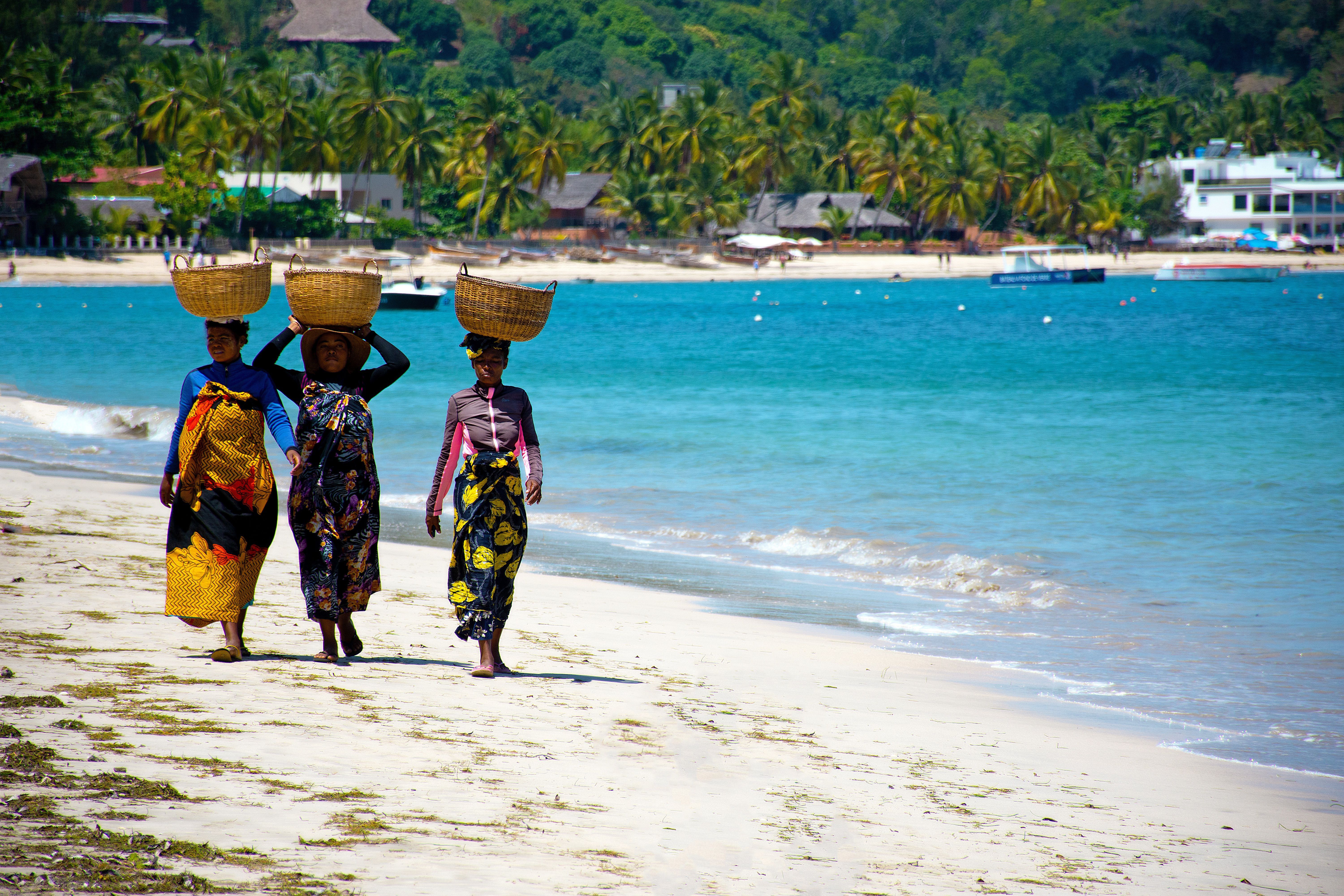 Tři ženy nesoucí koše na hlavách kráčejí po písečné pláži s palmami na Madagaskaru.