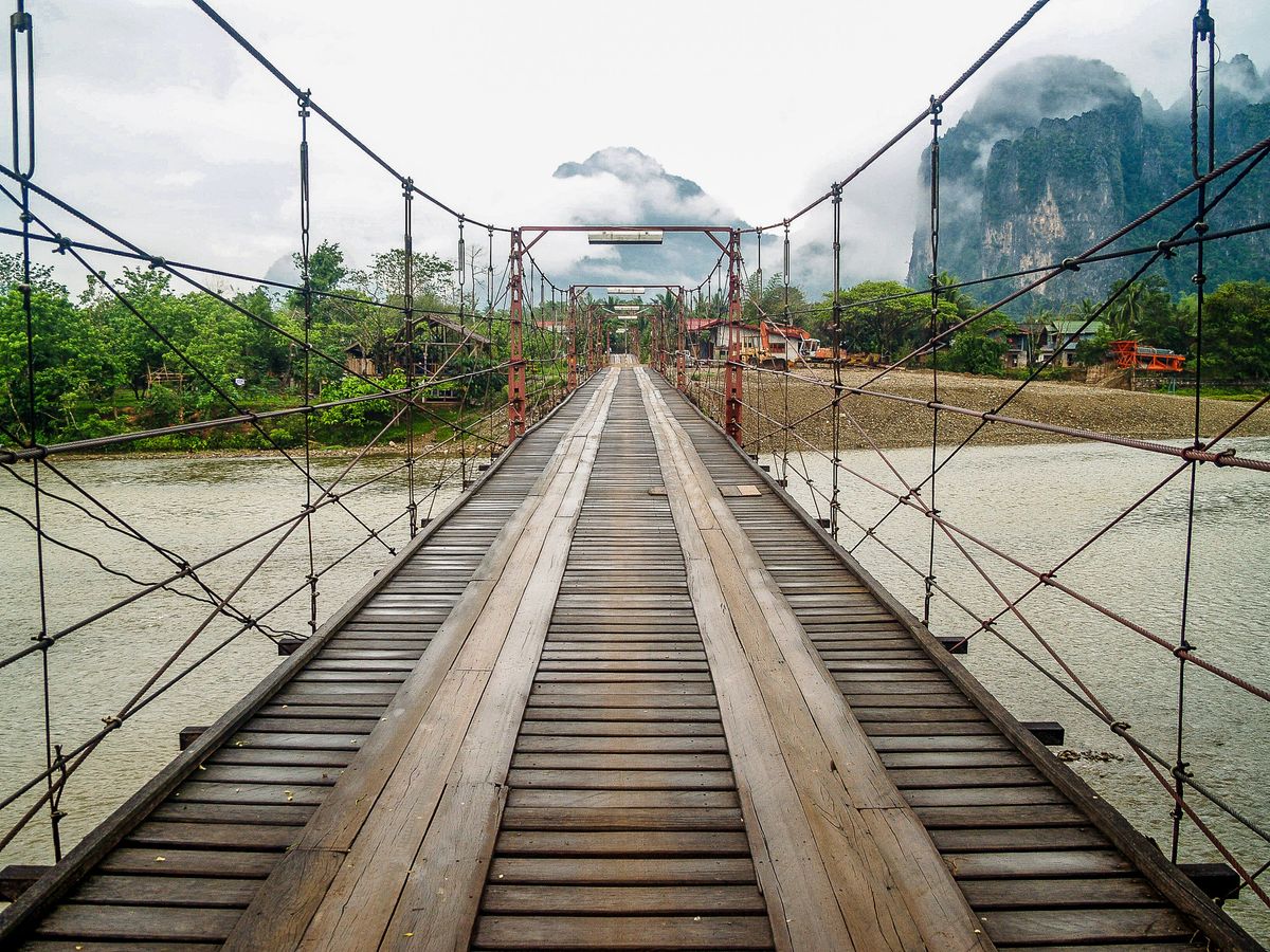 Dřevěný visutý most přes řeku v&nbsp;městečku Vang Vieng obklopený tropickou zelení a&nbsp;dramatickými vápencovými skalami.
