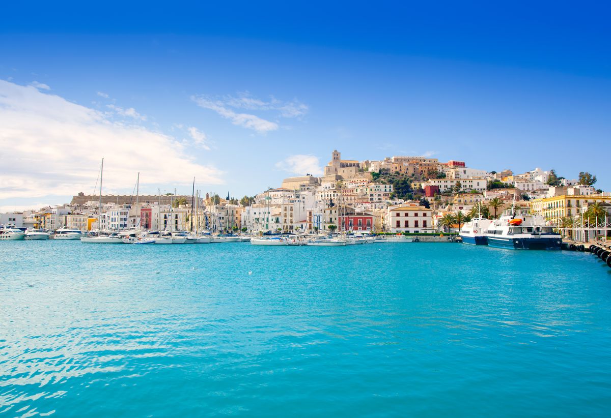 Pohled na historické centrum města Eivissa na ostrově Ibiza se starobylými budovami a přístavem plným jachet a lodí na azurovém moři.