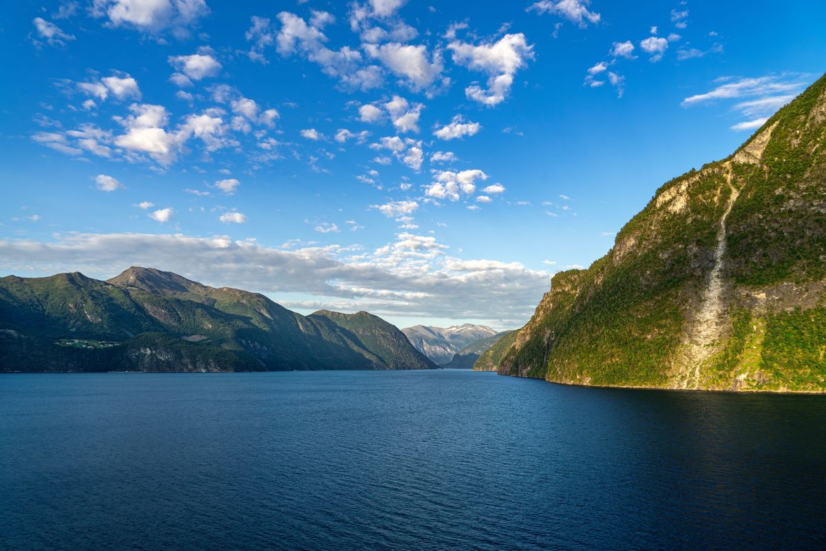 Pohled na norský fjord se strmými zalesněnými svahy a klidnou vodní hladinou pod modrou oblohou s bílými mraky.