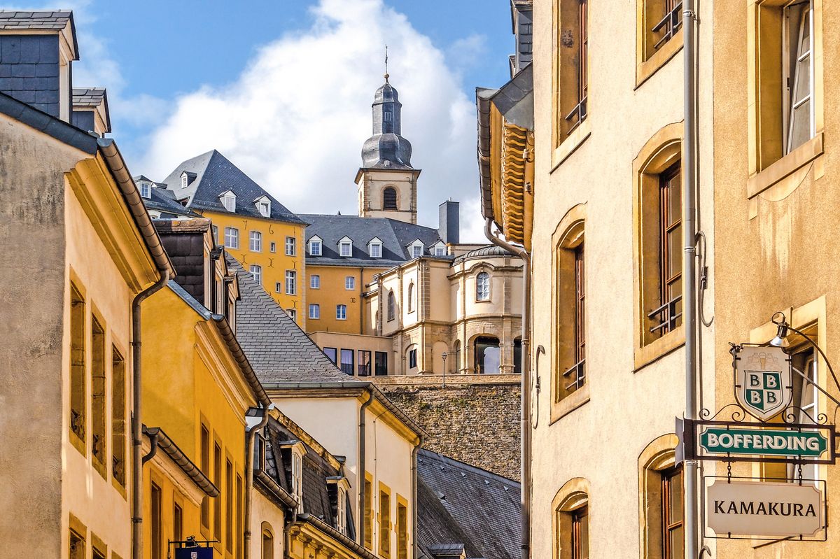 Úzká ulice s&nbsp;historickými budovami ve městě Lucemburk, kde pozadí dominuje věž kostela s&nbsp;typickou architekturou.