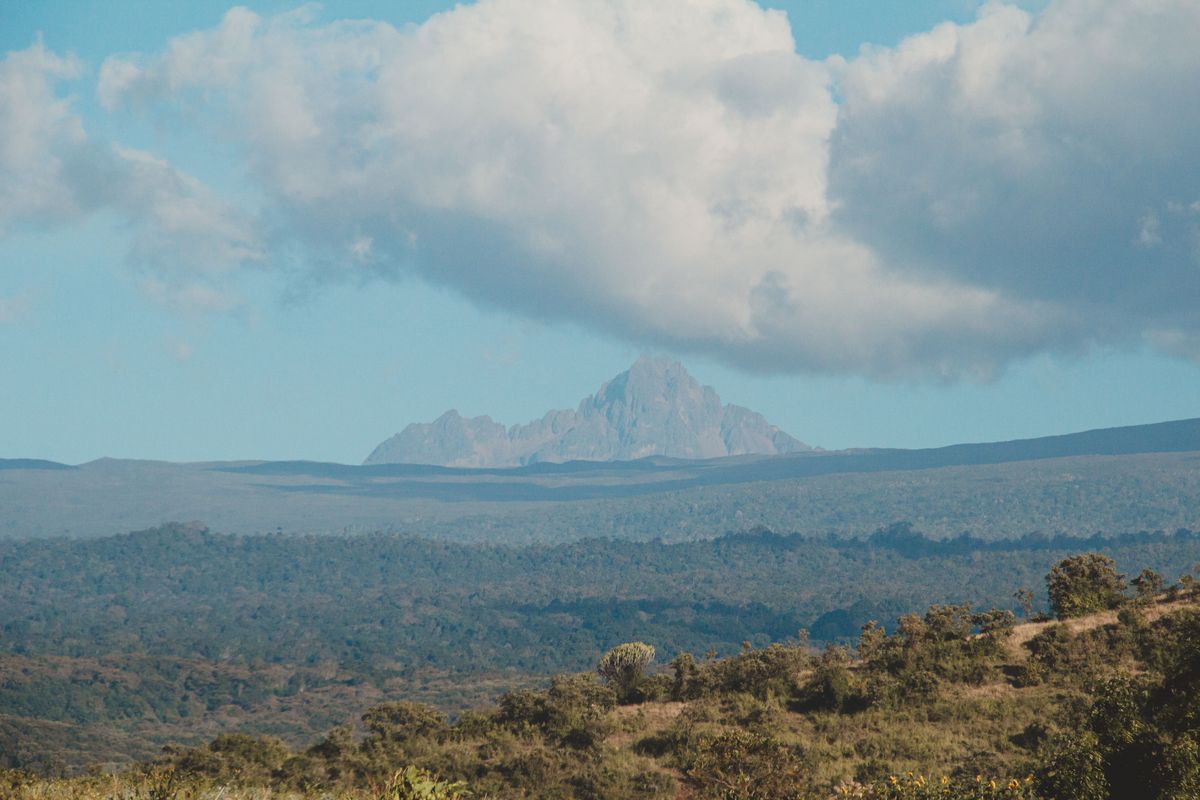 Horská scenerie s&nbsp;impozantním vrcholem Mount Kenya, kolem nějž se rozprostírají zelené lesnaté svahy a&nbsp;modrá obloha s&nbsp;oblačností.