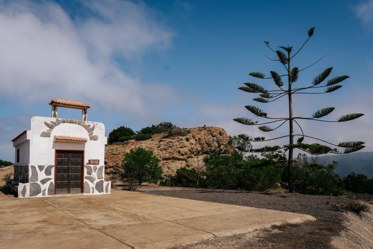 Malá bílá kaple stojí na okraji útesu na ostrově La Gomera obklopená bujnou vegetací a vysokým zvláštním stromem.