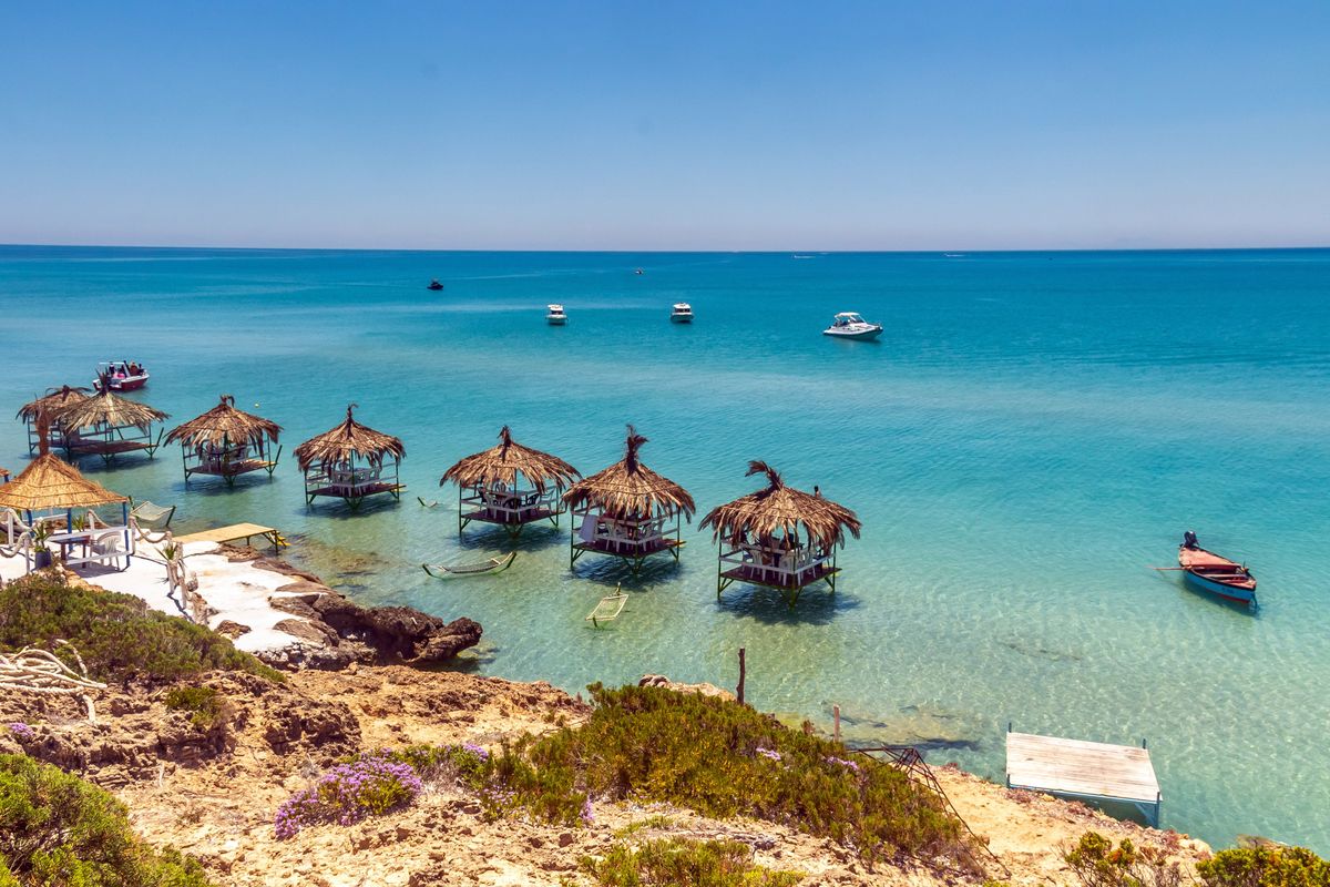 Tropické bungalovy s doškovými střechami stojí na písčité pláži ostrova Djerba a nabízejí výhled na klidné Středozemní moře.