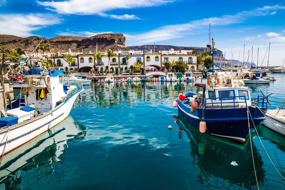 Přístav plný lodí obklopený malebnou architekturou v Puerto de Mogán na Gran Canarii s horami v pozadí.