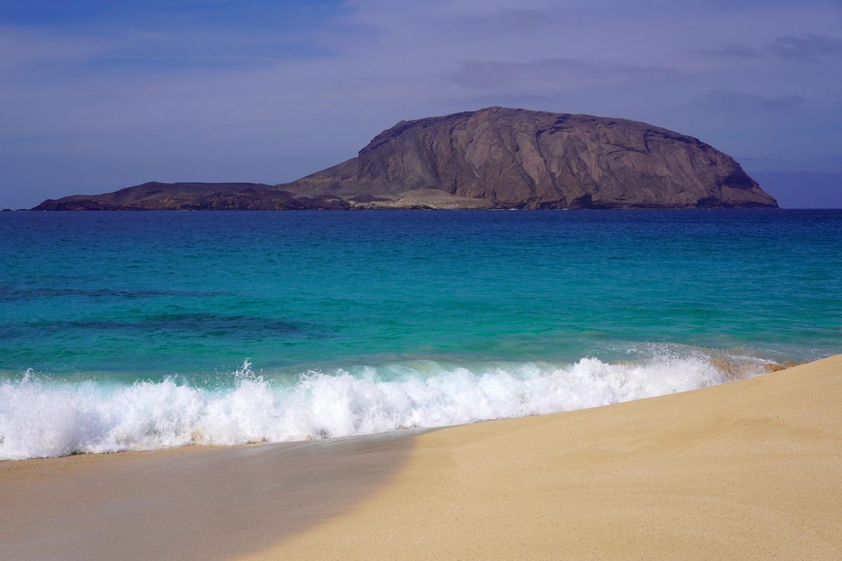Zlatavá písečná pláž s tyrkysovým mořem na ostrově Lanzarote s impozantním skalním útvarem na horizontu.
