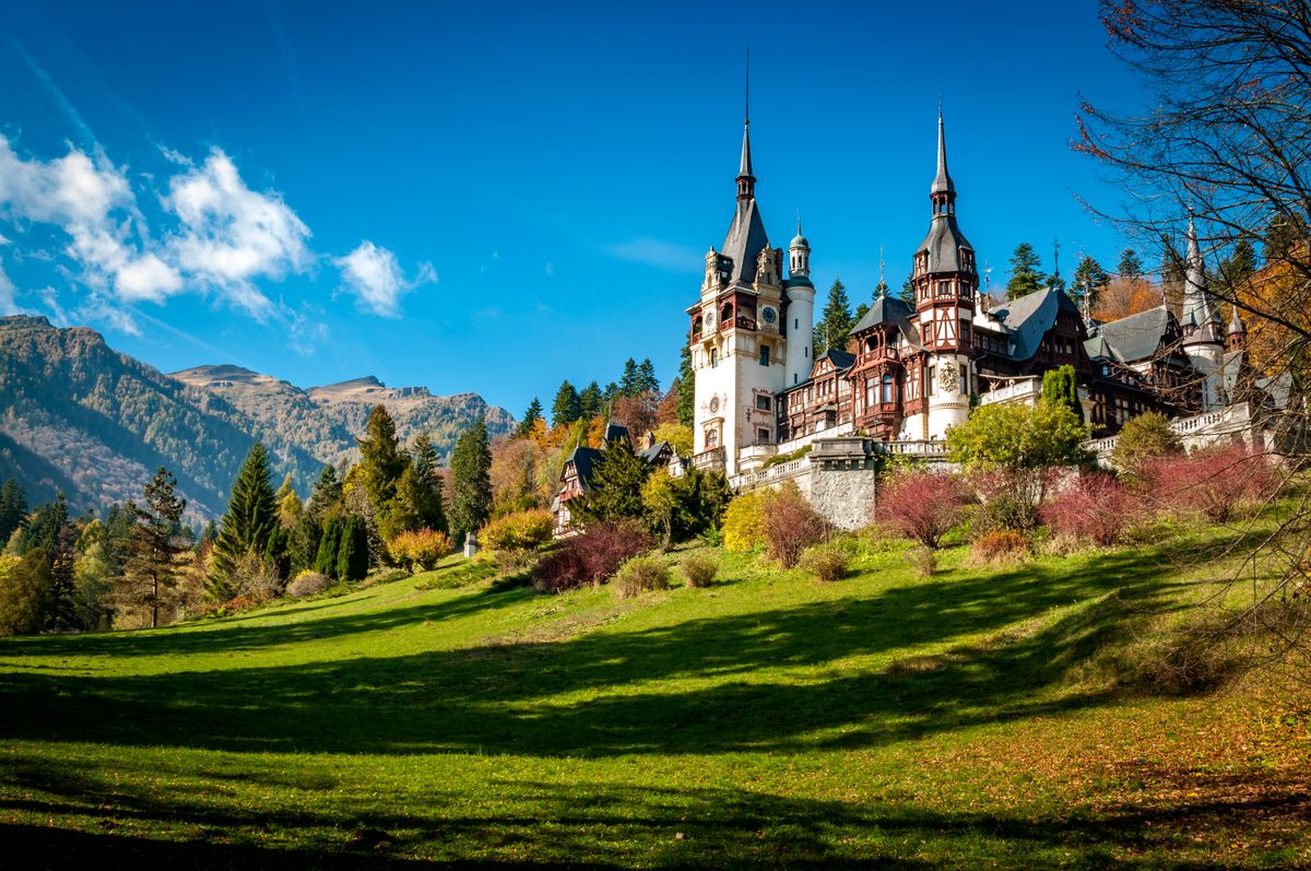 Pohled na zámek Peleș, obklopený horami a lesy v rumunské Sinii. Krásná přírodní scenérie s modrou oblohou doplňuje historickou architekturu.
