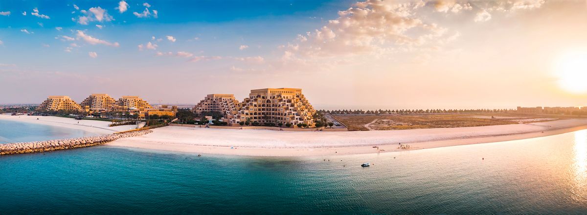 Panoramatický pohled na pláž v emirátu Ras al-Khaimah, kde se zlatavý písek setkává s modrou vodou moře, a v pozadí jsou vidět moderní hotelové komplexy.