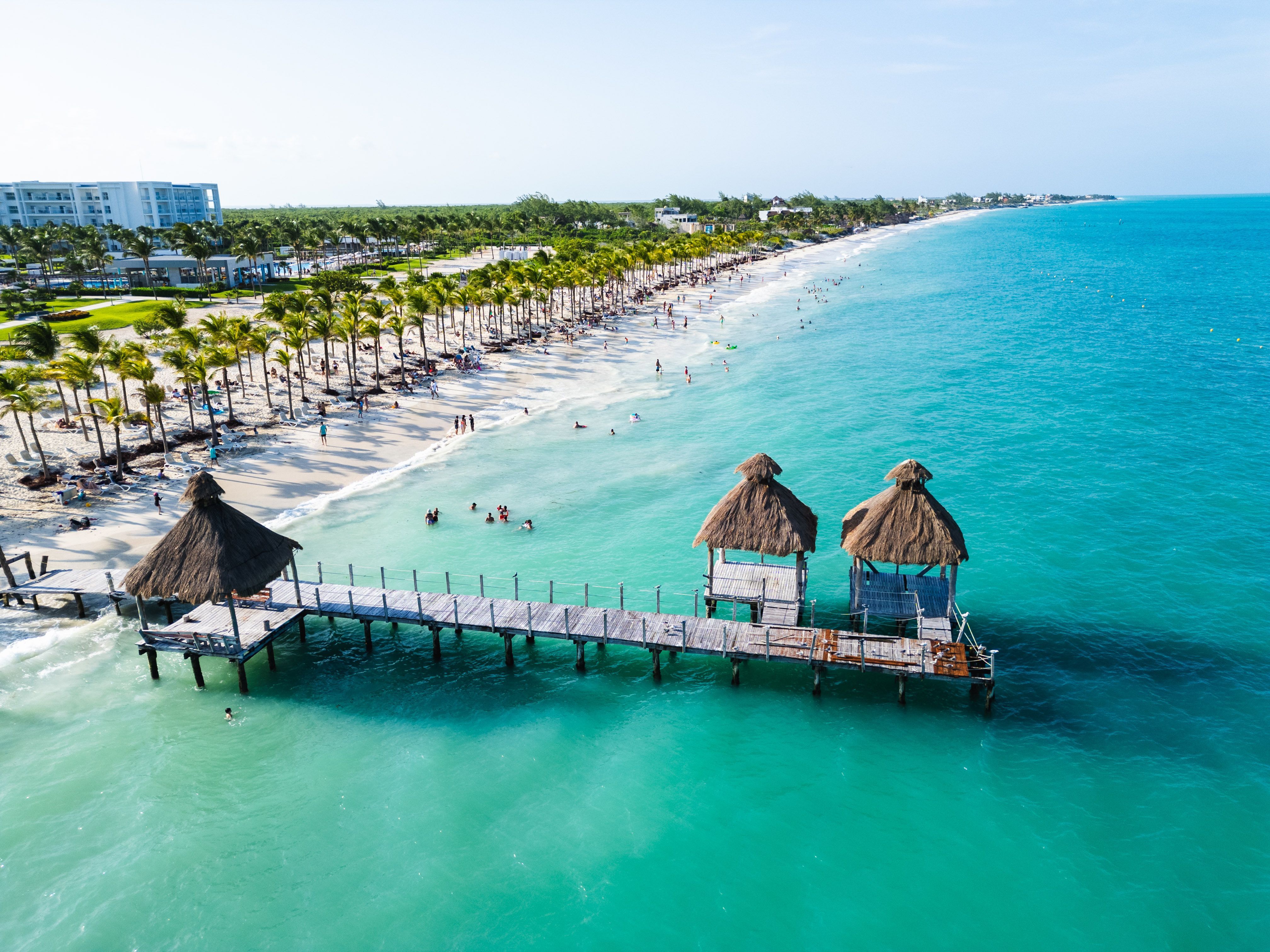 Dřevěné molo s&nbsp;palmovou střechou vede nad azurovým mořem k&nbsp;bílému písku Playa del Carmen v&nbsp;Mexiku, které lemují palmy a&nbsp;odpočívající lidé.