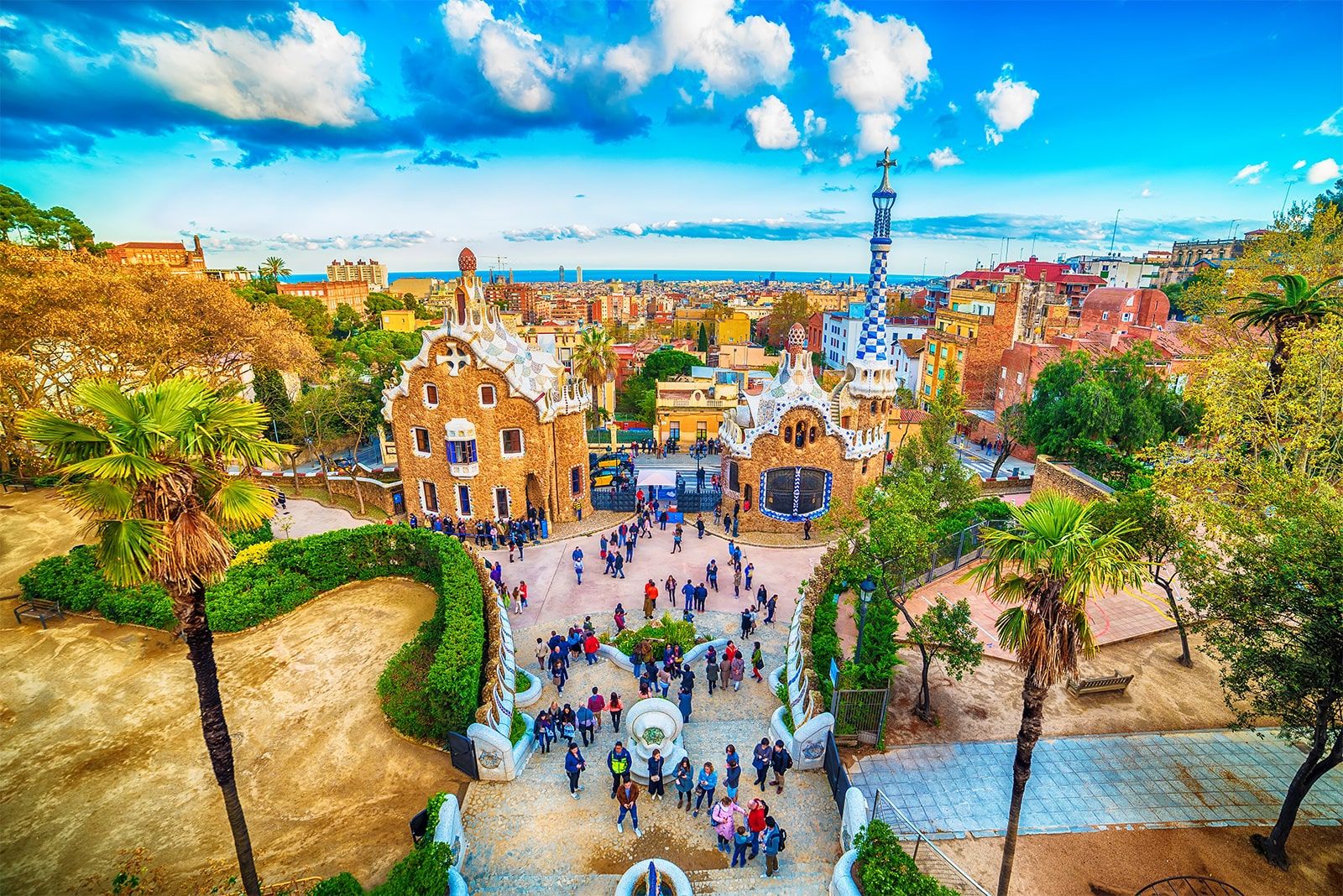 Park Güell v Barceloně zobrazuje ikonické barevné budovy oživené mozaikou, obklopené zelení a palmami, s výhledem na město a moře.