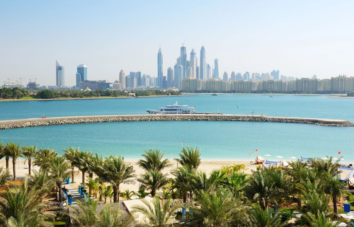 Pohled na dubajské panorama s ikonickými mrakodrapy na pozadí a pláží s palmami v popředí.