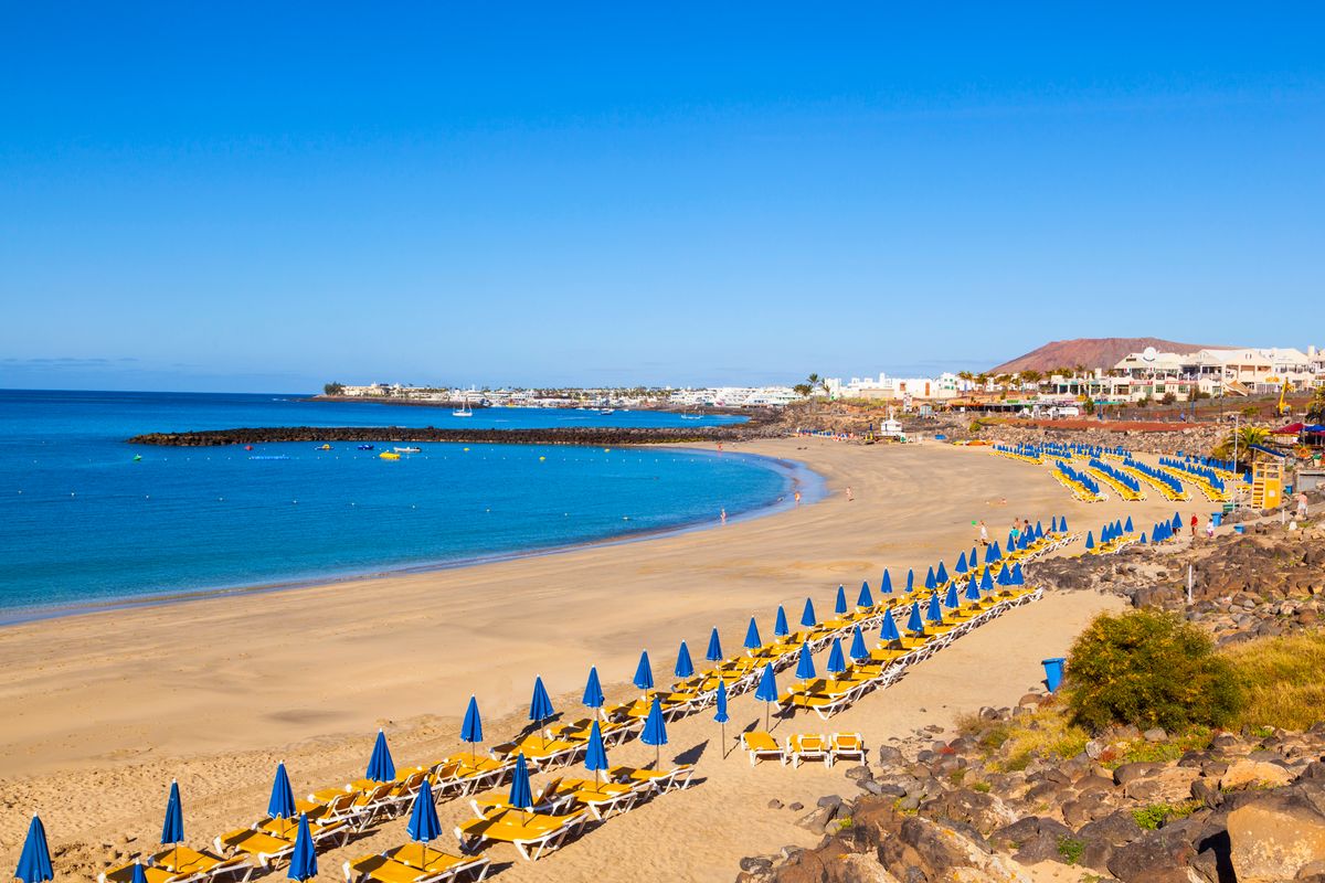 Pláž s modrými slunečníky a lehátky na ostrově Lanzarote s výhledem na modré moře a místní budovy v pozadí.