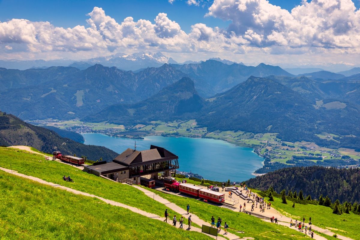 Scenérie alpského jezera Wolfgangsee s obklopenými horami a horskou železnicí v popředí.
