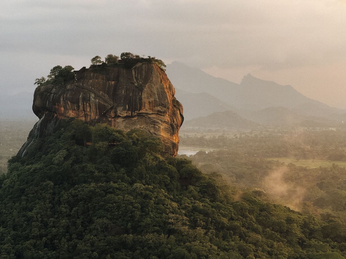 Skalní pevnost Sigiriya na Srí Lance se tyčí nad okolní hustou zelení za jasného dne s dramatickými horami na pozadí.