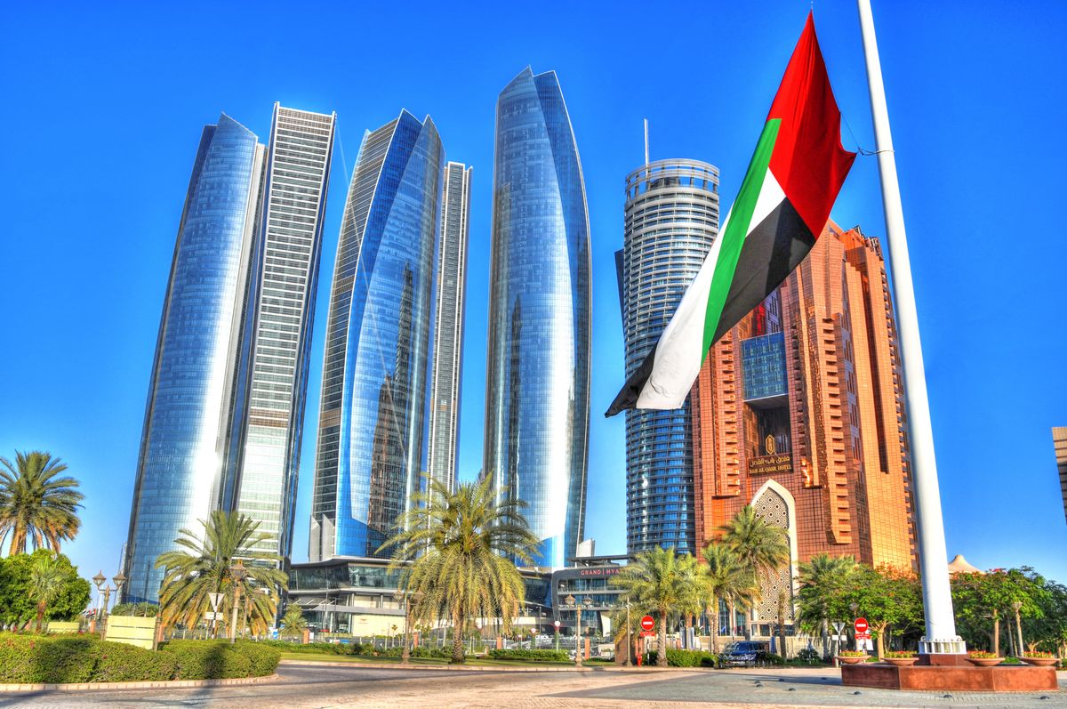 Moderní mrakodrapy Etihad Towers v Abú Dhabí obklopené palmami a vlající vlajka Spojených arabských emirátů.