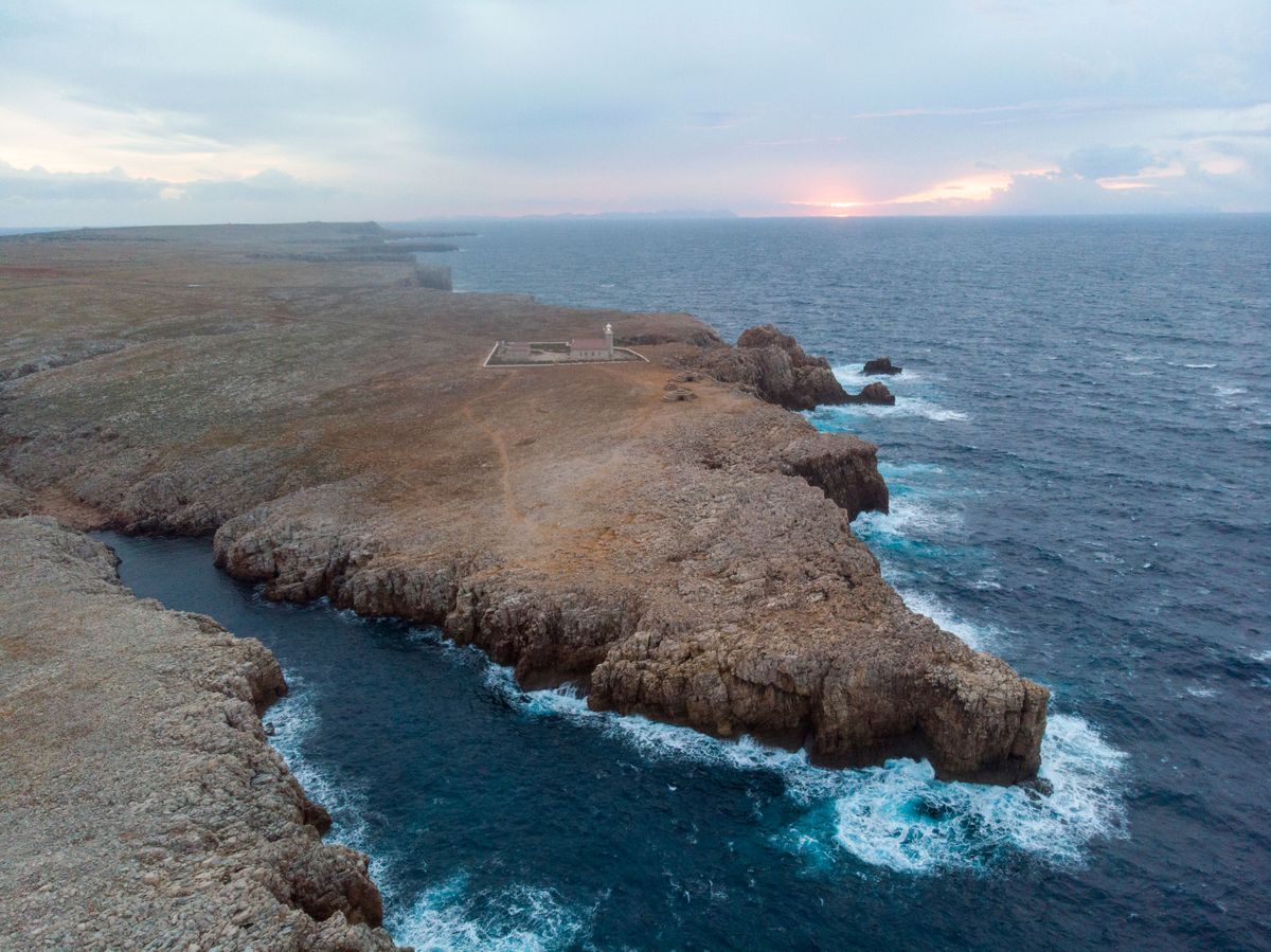 Pobřeží ostrova Menorca s majákem na skalnatém výběžku a rozbouřeným Středozemním mořem při západu slunce.