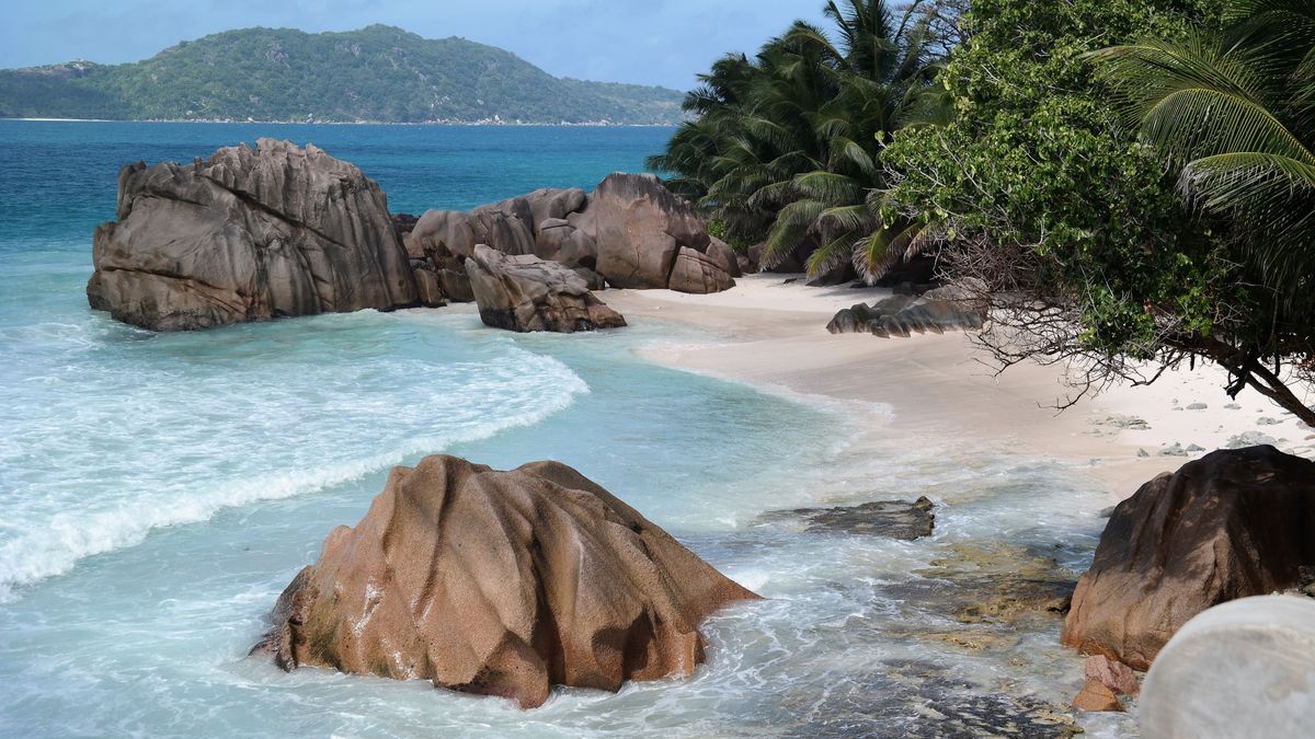 Tropická pláž s jemným bílým pískem, velkými žulovými kameny a palmami obklopená azurovým oceánem na Seychelách.