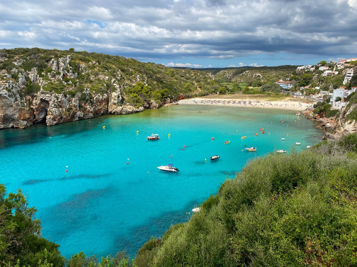 Zátoka na ostrově Menorca ve Španělsku s tyrkysově modrým mořem, pláží obsazenou slunečníky a okolními skalami porostlými vegetací.