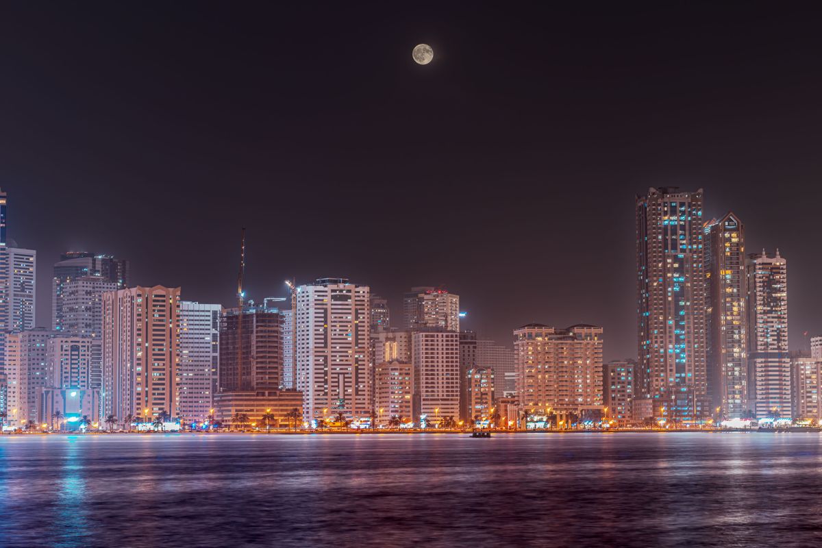 Panorama nočního města Ajman v Spojených arabských emirátech s osvětlenými mrakodrapy a měsícem na obloze.