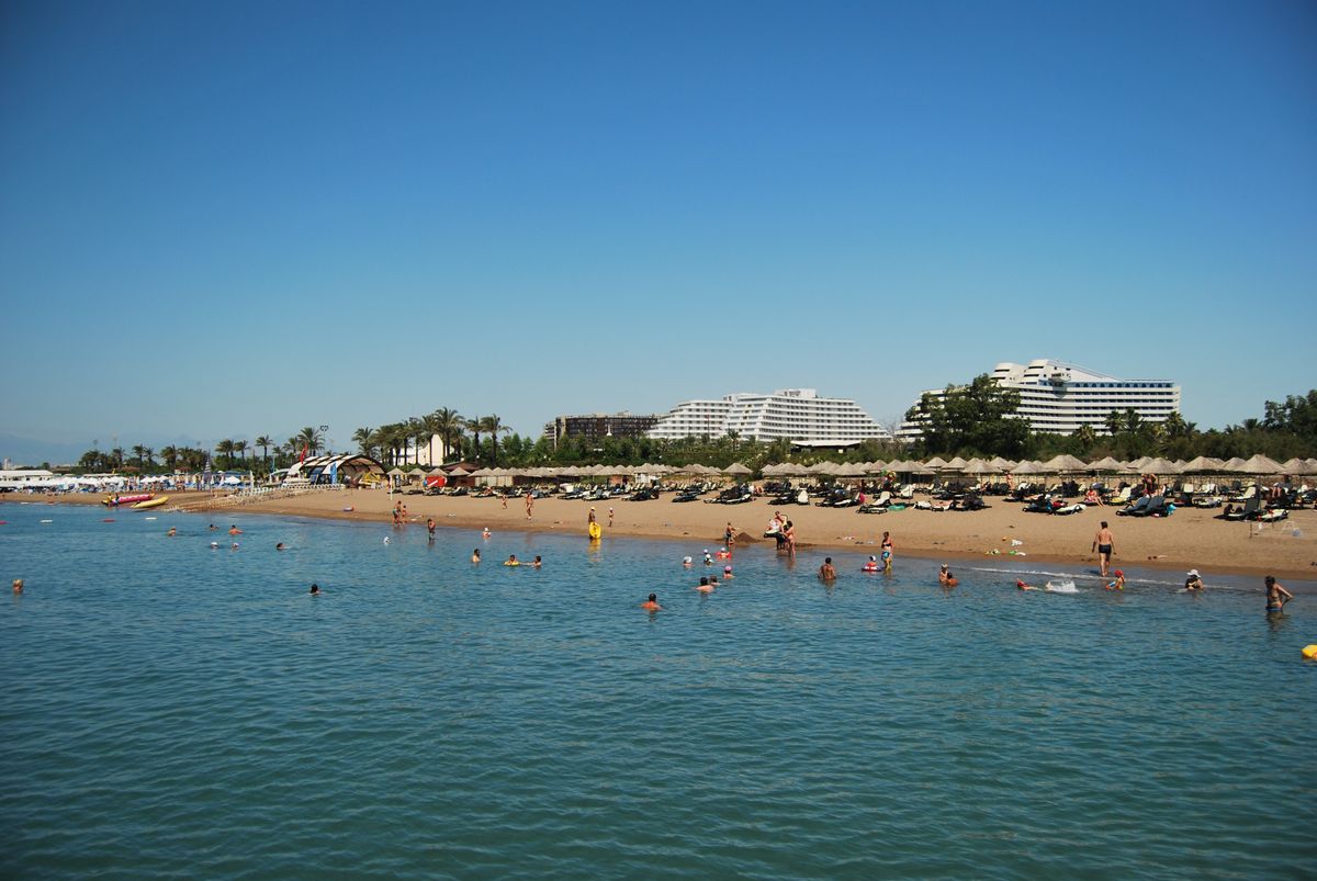 Pláž s čistým mořem a turisté využívající lehátka v oblasti Belek na Turecké riviéře, za nimi moderní hotelové komplexy.