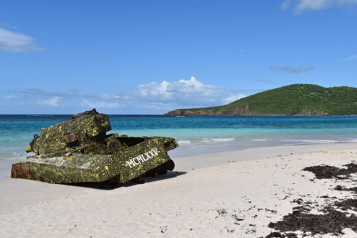 Starý vojenský tank pokrytý pestrobarevnými vzory na břehu pláže Playa Flamenco s&nbsp;tyrkysovým mořem v&nbsp;pozadí.