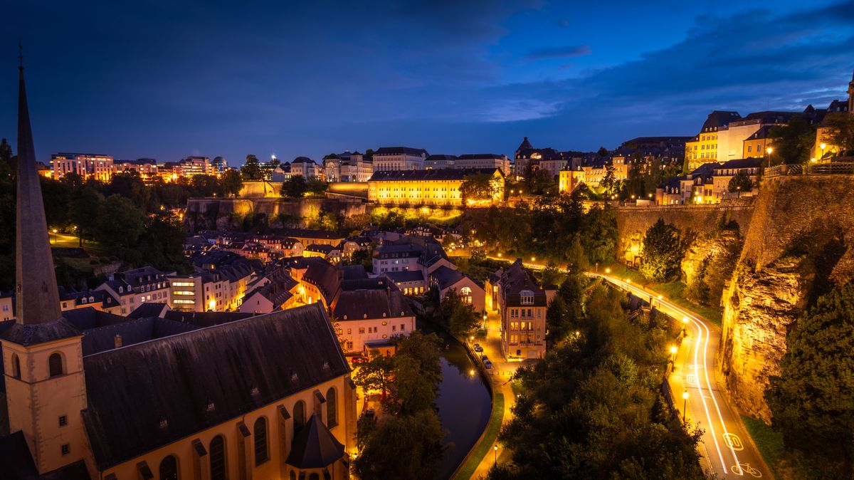 Noční pohled na osvětlené centrum města Lucemburk s&nbsp;historickou architekturou a&nbsp;klikatící se řekou Alzette.